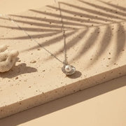 Necklace with a shell and pearl pendant on a sandy surface with a palm leaf shadow.