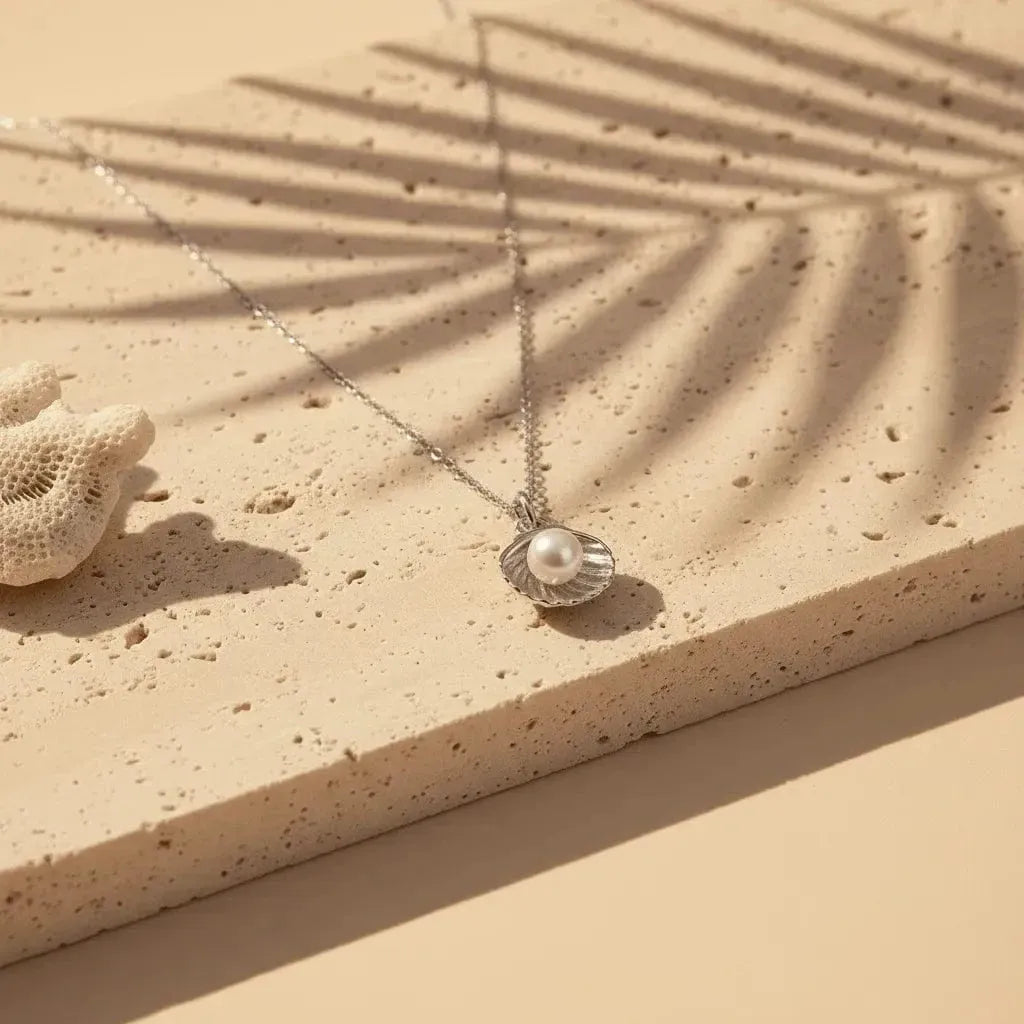 Necklace with a shell and pearl pendant on a sandy surface with a palm leaf shadow.