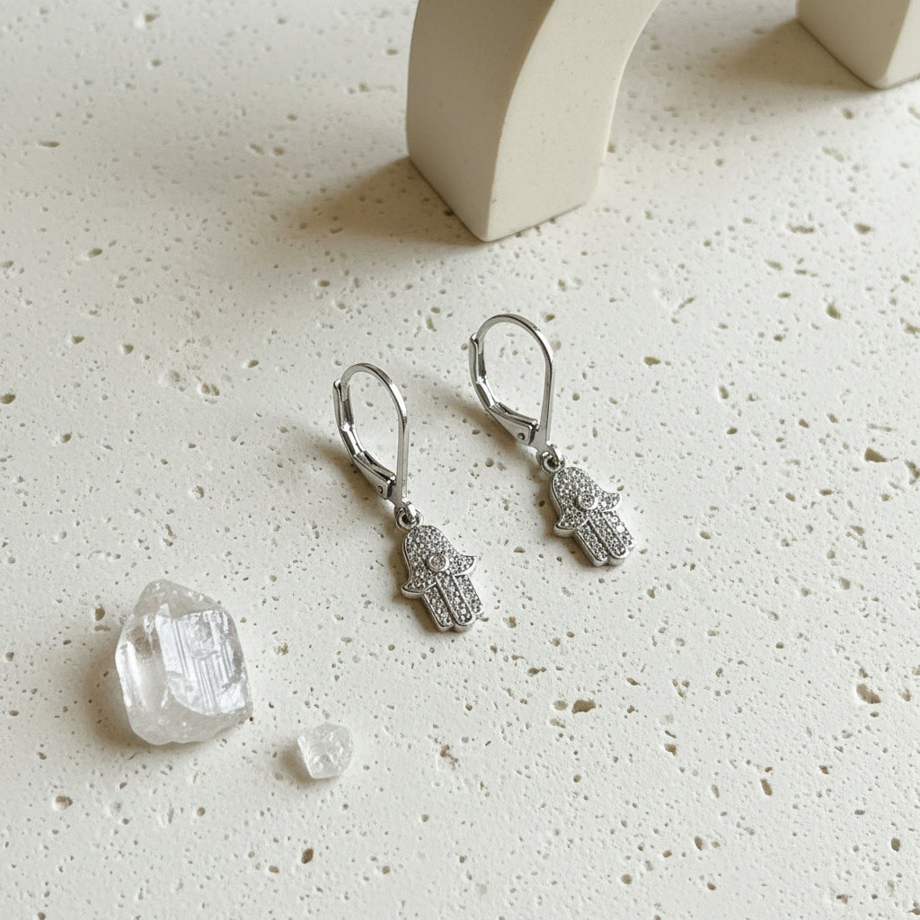 Silver plated crystal hamsa hand earrings with eye detail on leverbacks, on white stone