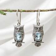 Silver vintage owl drop earrings with blue crystal accent stones, hanging from a branch
