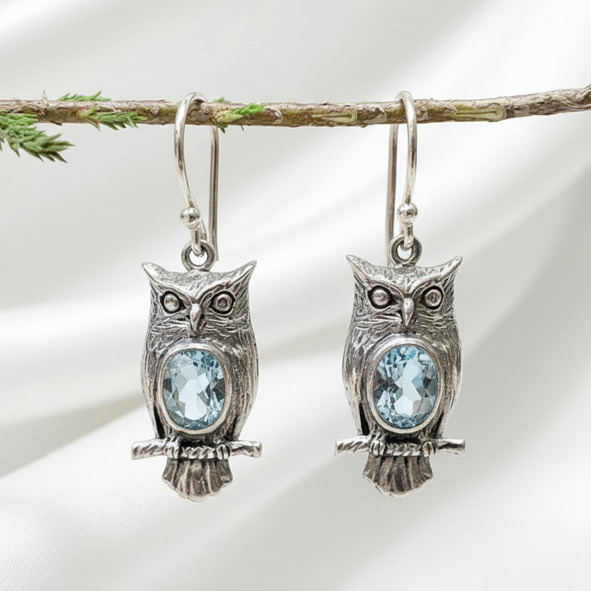 Silver vintage owl drop earrings with blue crystal accent stones, hanging from a branch