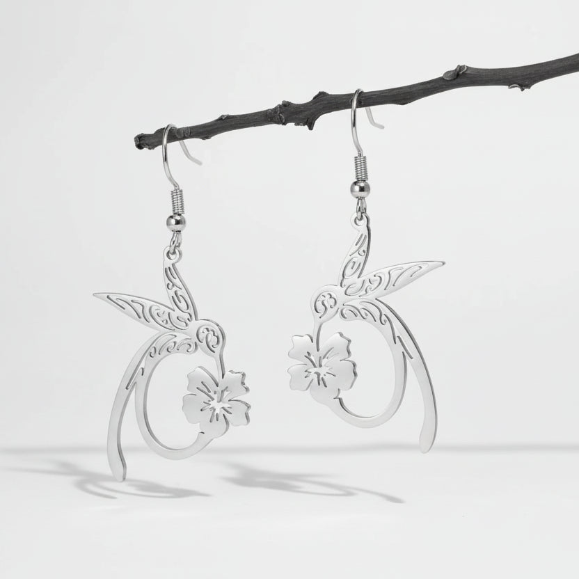 Silver hummingbird and floral earrings hanging on a branch against a dark green background