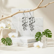 Silver orchid earrings on a marble stand with white orchids and green leaves in the background.