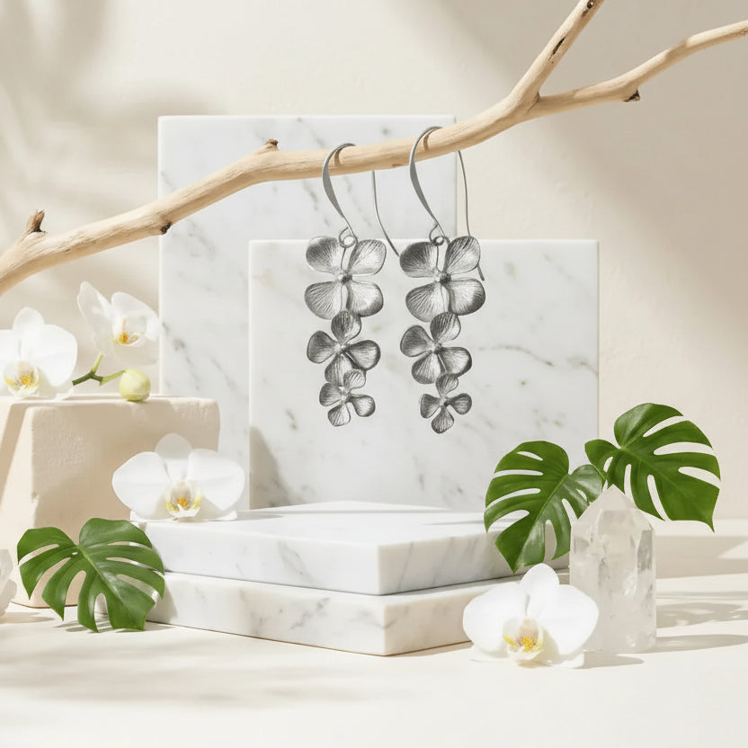 Silver orchid earrings on a marble stand with white orchids and green leaves in the background.