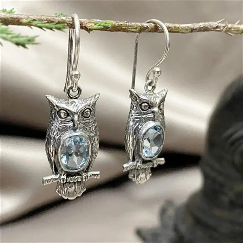 Silver vintage owl drop earrings with blue crystal accent stones, hanging from a branch