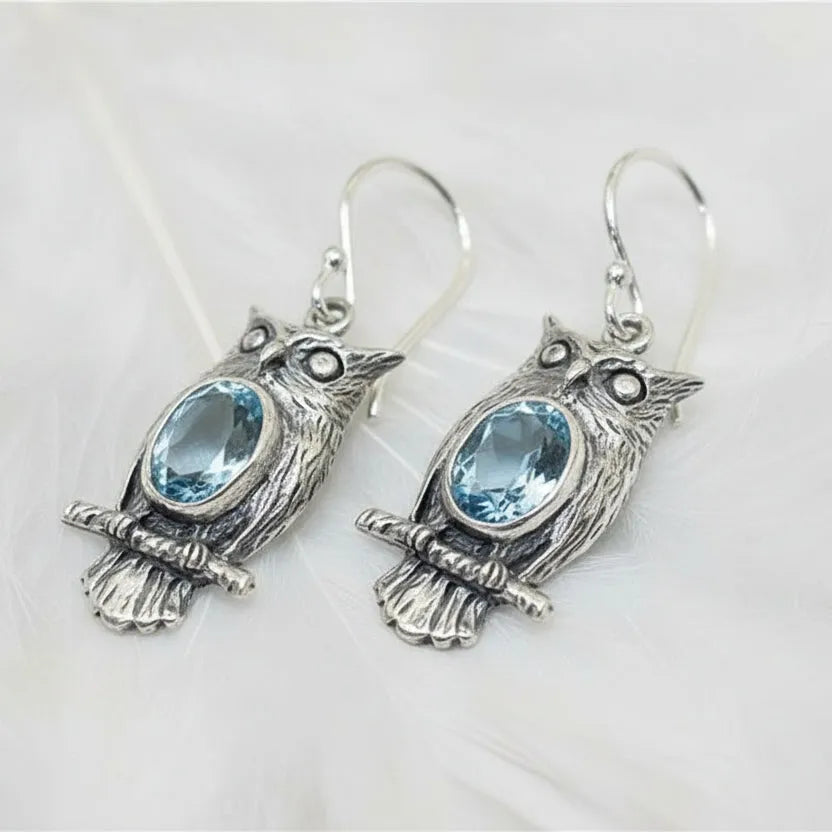 Silver vintage owl drop earrings with blue crystal accent stones, hanging from a branch