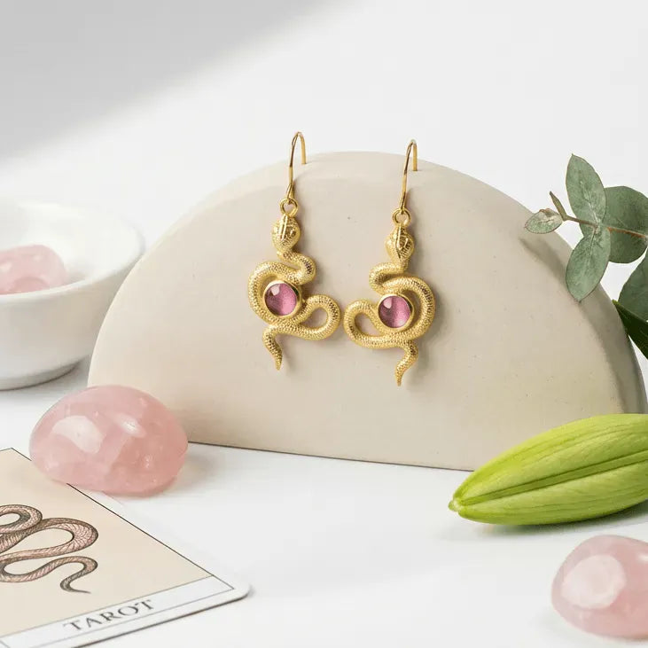 Gold earrings with pink stones on a white surface with crystals and a card.