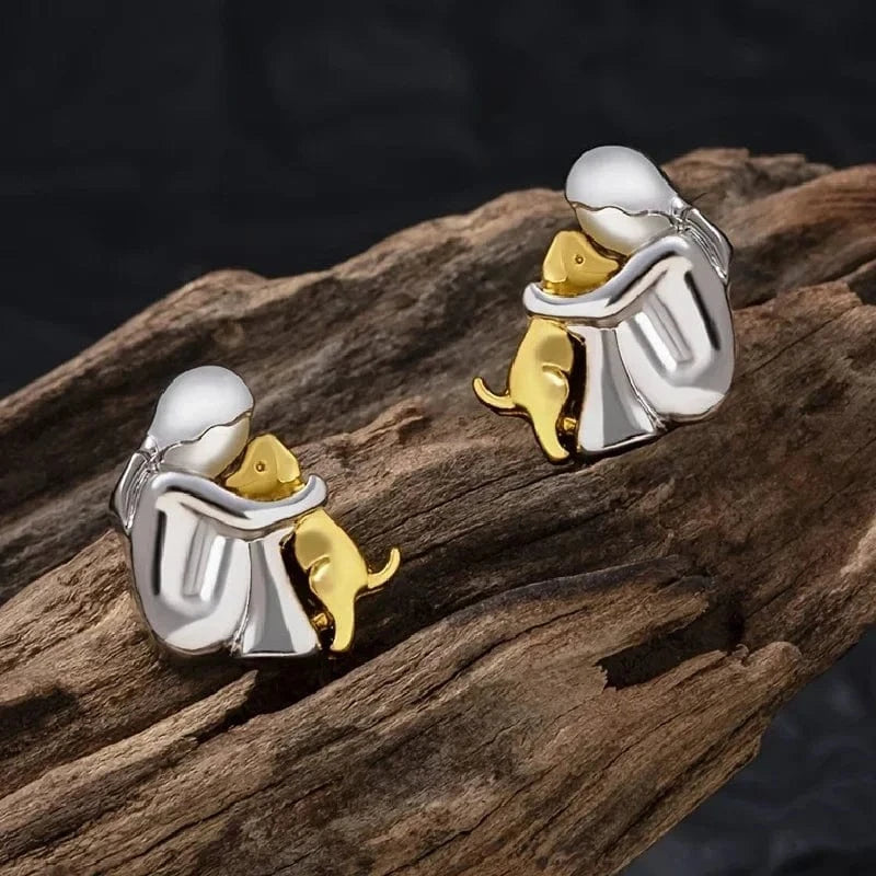 Silver and gold earrings shaped like a person hugging a dog on a wooden surface