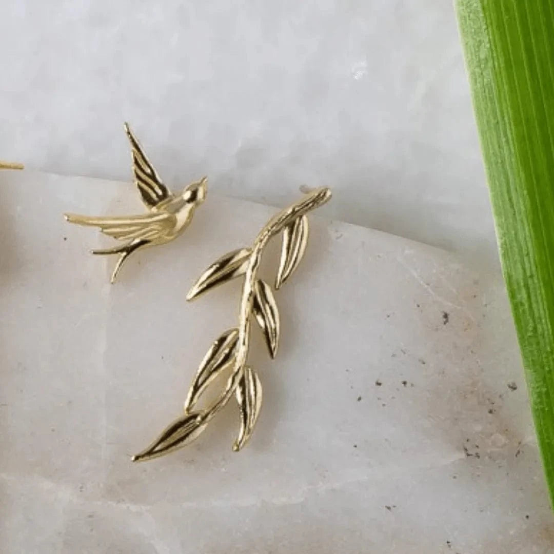 Gold bird and leaf earrings on a white stone surface with a green leaf in the background