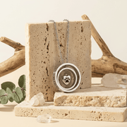 Necklace with a paw print pendant on a natural stone stand with driftwood and eucalyptus leaves in the background.