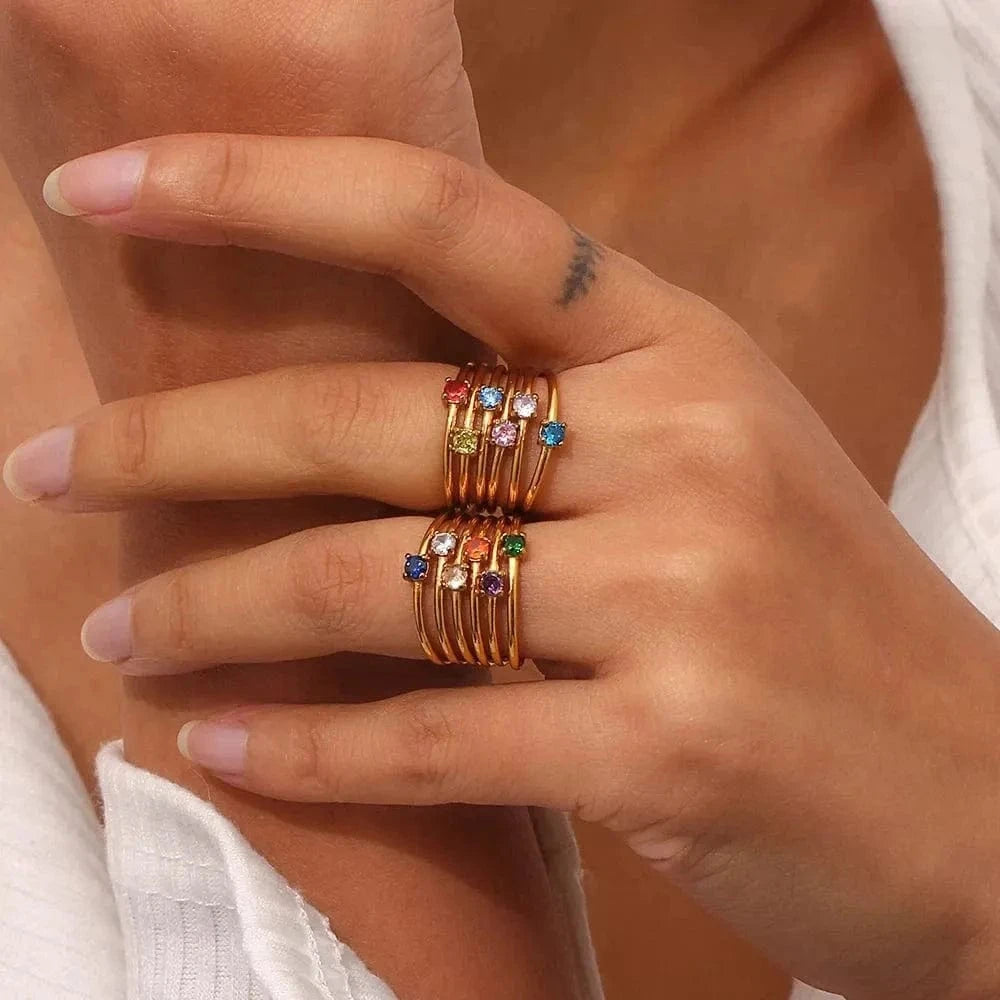 Hand wearing multiple gold rings with colorful gemstones on a neutral background