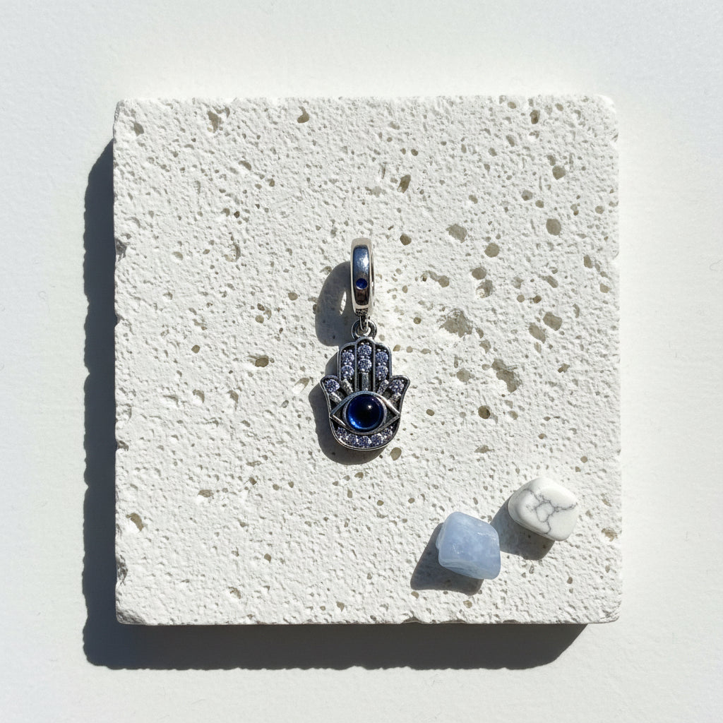 Sterling silver hamsa hand pendant with a deep blue eye and pavé crystals, on white stone beside blue and white crystals