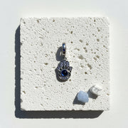 Sterling silver hamsa hand pendant with a deep blue eye and pavé crystals, on white stone beside blue and white crystals