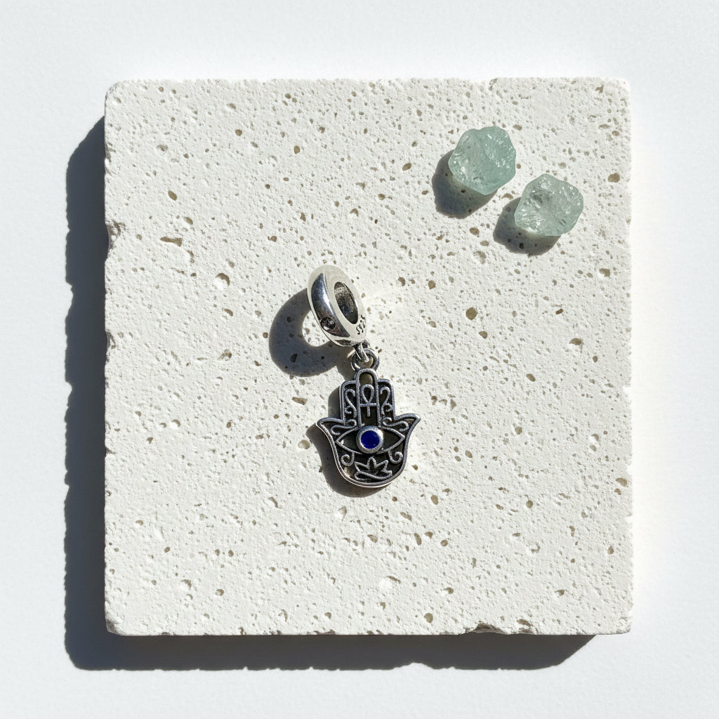 Cut-out sterling silver hamsa hand pendant with a small blue eye, on white stone beside green crystals