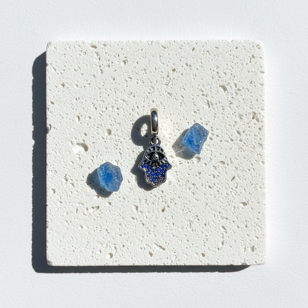 Double layered sterling silver hamsa hand pendant with a cut-out silver overlay, on white stone beside blue crystals
