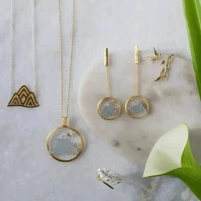 Gold jewelry set including necklaces, earrings, and rings on a marble surface with green leaves.