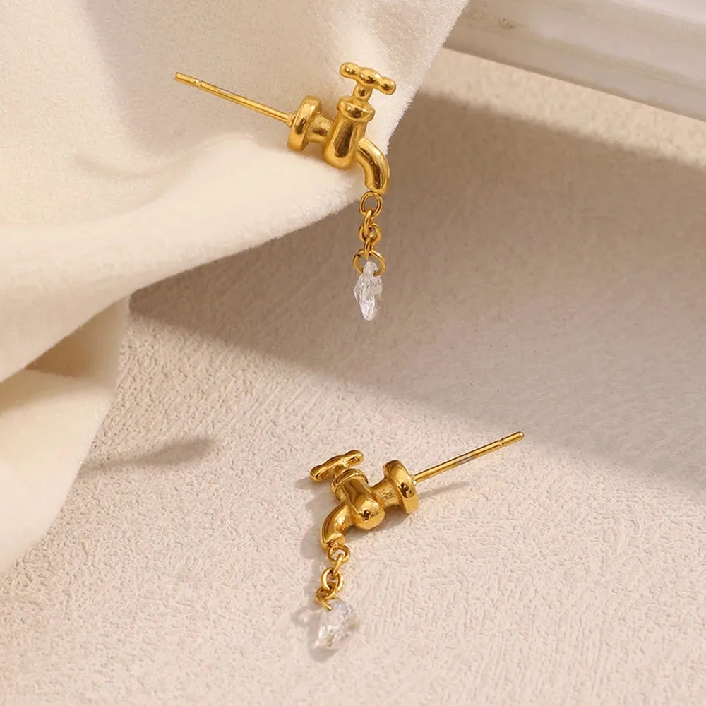 Gold faucet-shaped earrings with a small pearl on a beige surface