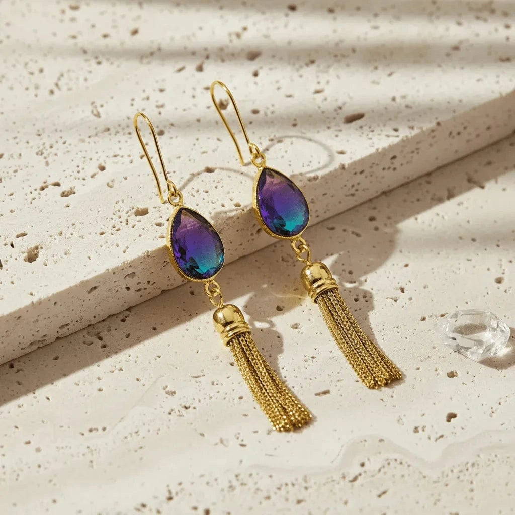 Gold earrings with purple teardrop stones and tassels on a textured beige surface