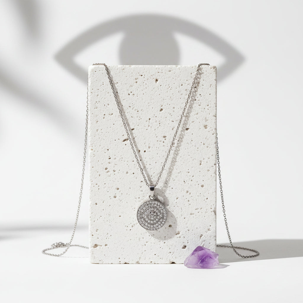 Silver third eye pavé necklace on pale stone, an eye-shaped shadow cast on the wall behind it.