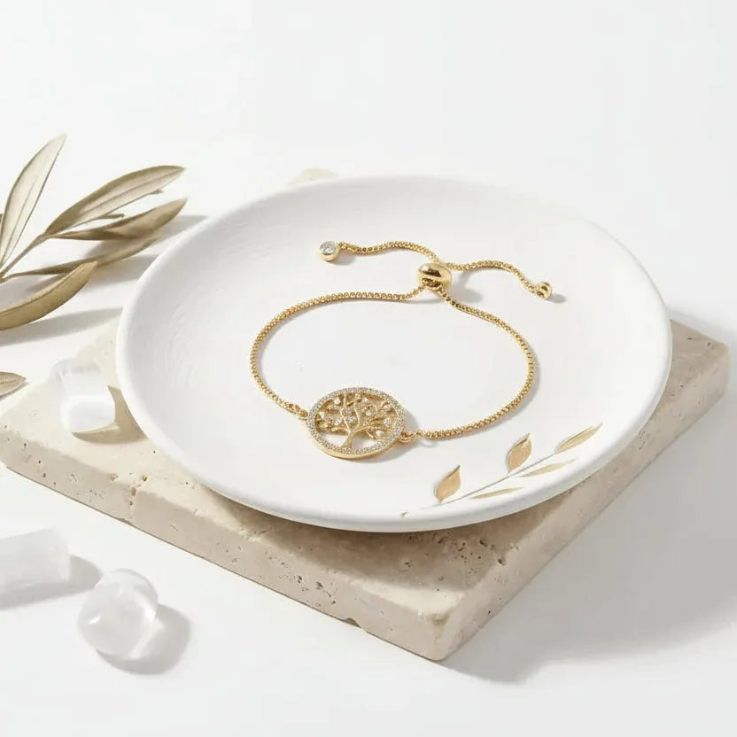 Gold Tree of Life adjustable bracelet on a white dish with leaves, on a light gray background.
