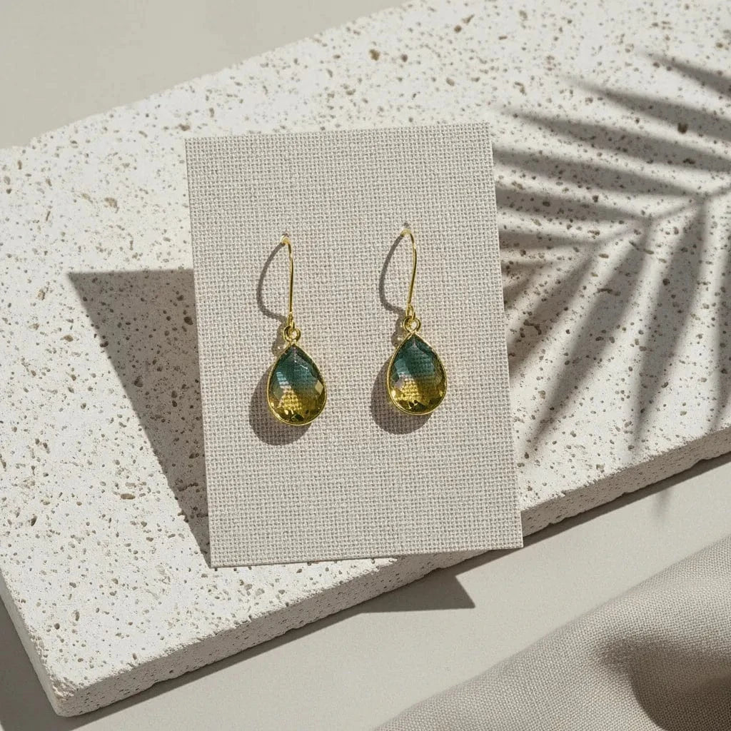 Gold earrings with green teardrop stones on a white card, placed on a textured stone surface.