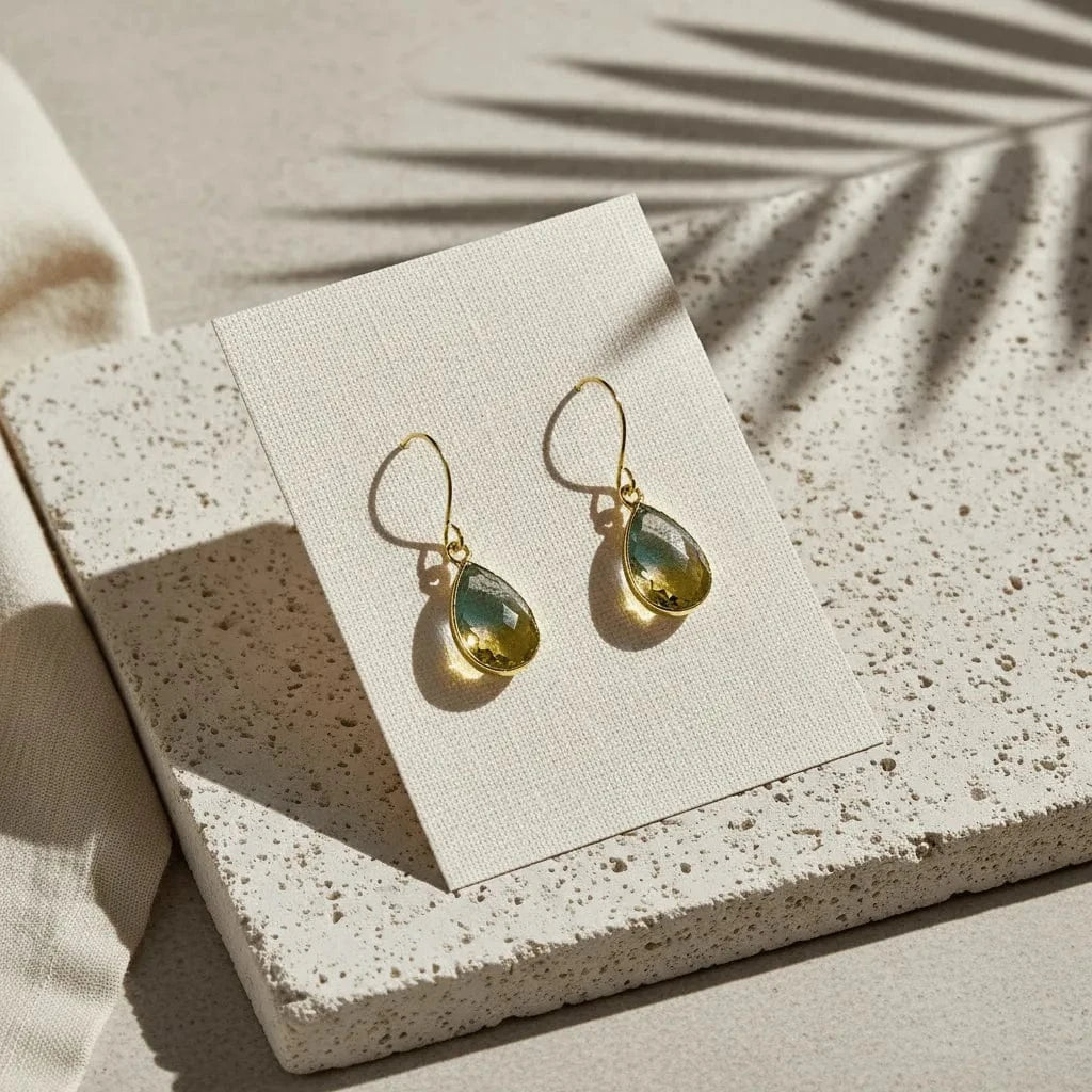 Gold earrings with green teardrop stones on a beige card, placed on a textured surface.