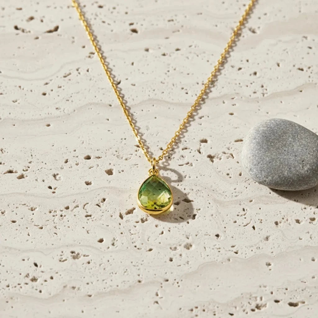 Gold necklace with a green teardrop pendant on a textured beige surface.