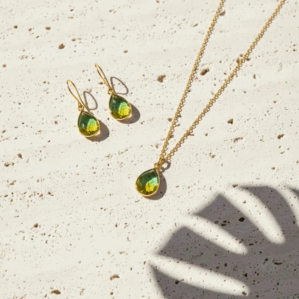 Gold necklace and earrings with green teardrop-shaped stones on a textured beige surface.