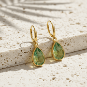 Gold earrings with green teardrop stones on a textured stone surface