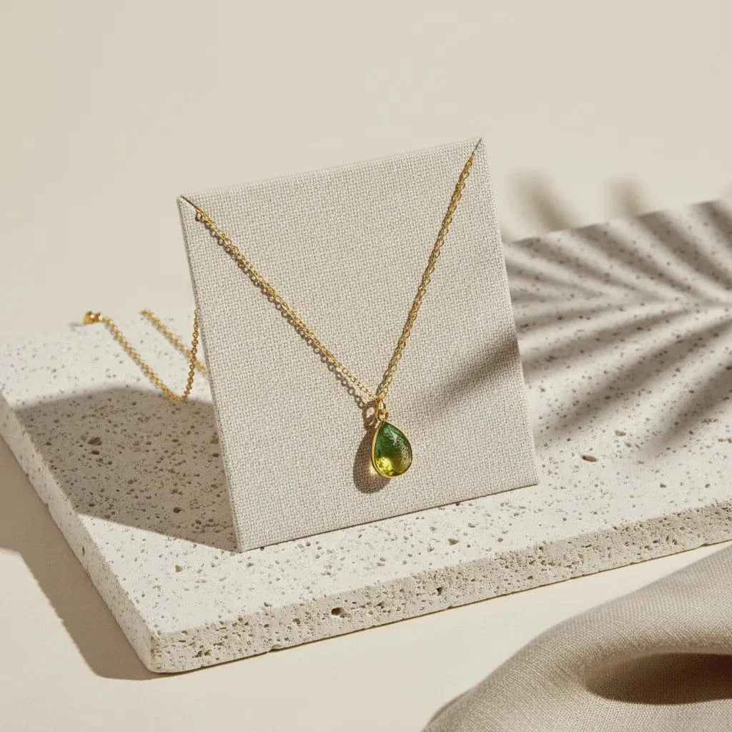 Gold necklace with a green teardrop pendant on a beige card, placed on a textured surface.