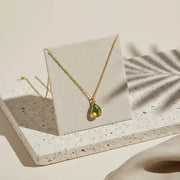Gold necklace with a green teardrop pendant on a beige card, placed on a textured surface.