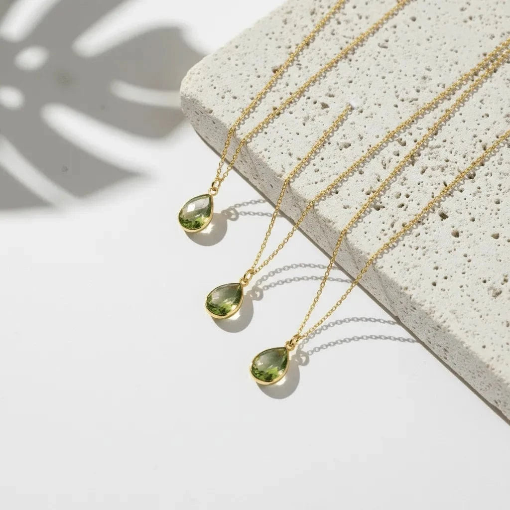 Three gold necklaces with green teardrop pendants on a light stone surface.