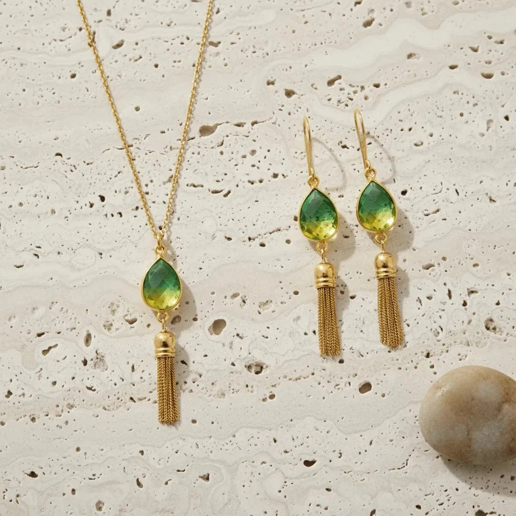 Gold necklace and earrings with green teardrop pendants on a textured beige surface.