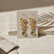 Gold floral earrings with green drops on a beige card against a textured beige background