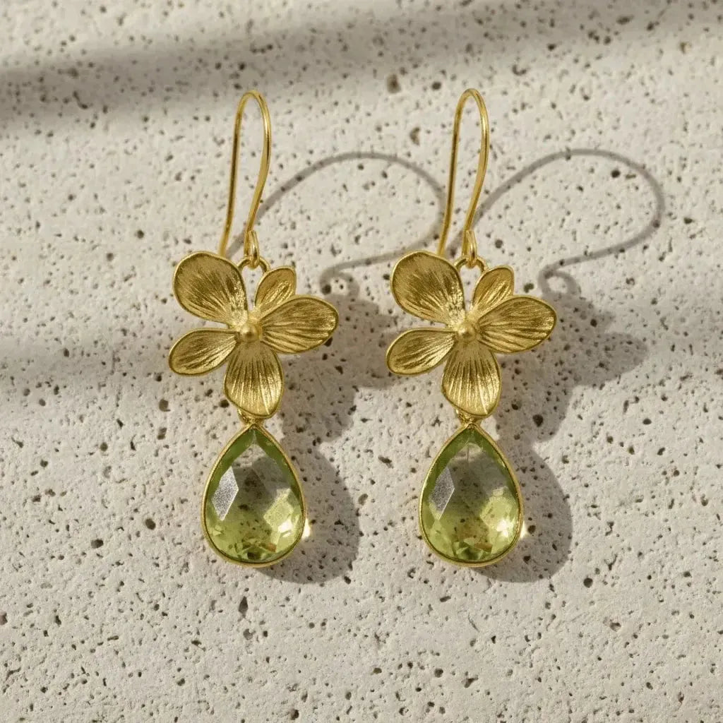 Gold flower-shaped earrings with green teardrop stones on a textured beige surface