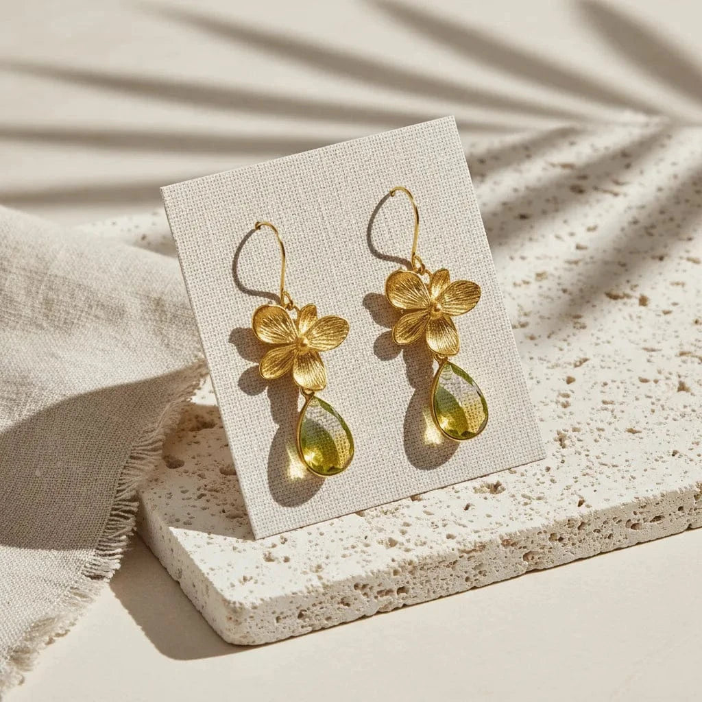 Gold flower earrings with green teardrop stones on a white card, placed on a textured beige surface.