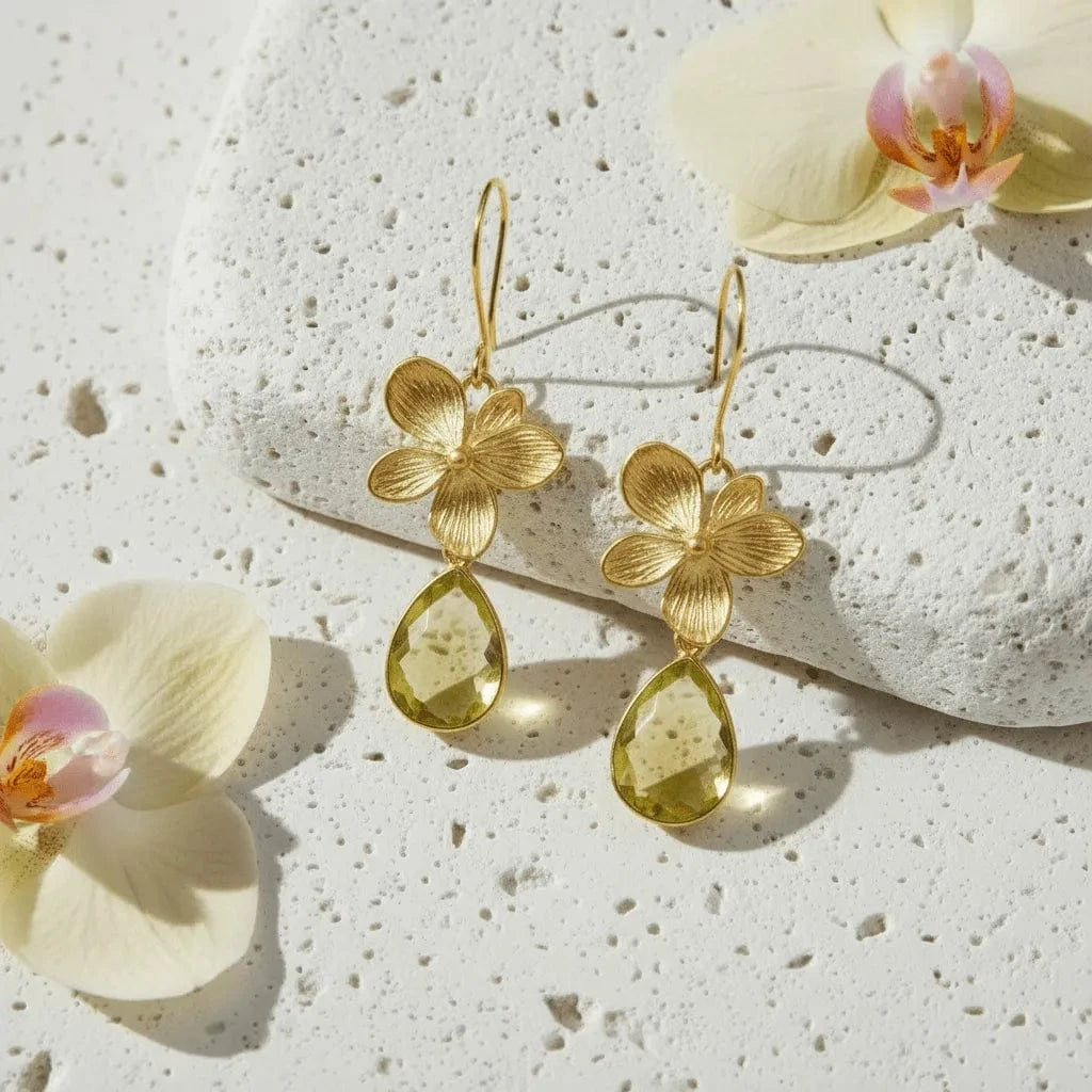 Gold flower earrings with green teardrop stones on a textured surface with flowers.