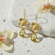 Gold flower earrings with green teardrop stones on a textured surface with flowers.