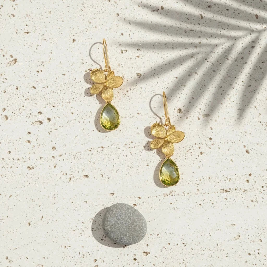 Gold earrings with floral designs and green teardrop stones on a textured surface.