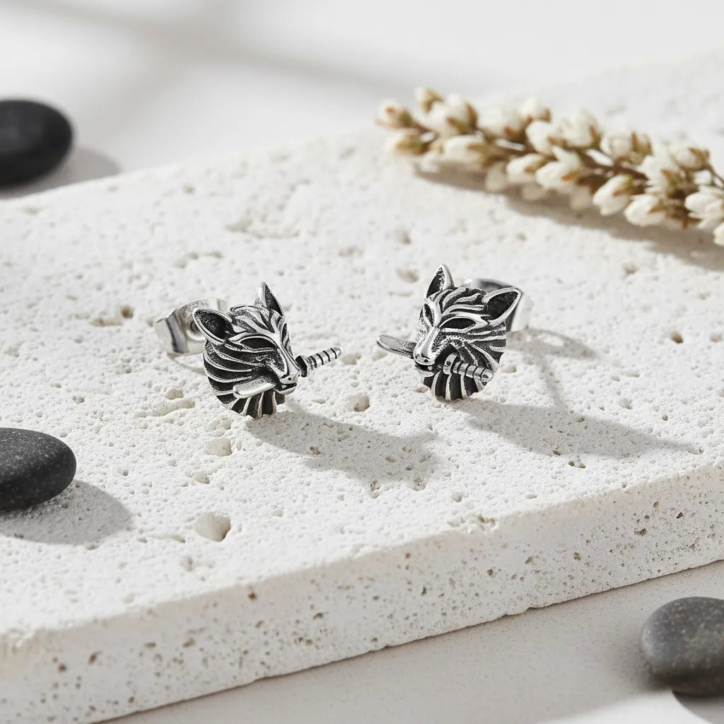 Silver wolf head earrings on a textured white surface with stones and dried plants.
